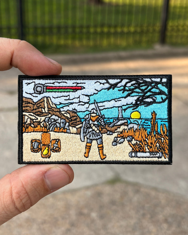 A rectangular embroidered patch held by a hand against a blurred outdoor background. The patch features a fantasy scene with a warrior, mountains, a sword, and a bright yellow sun.