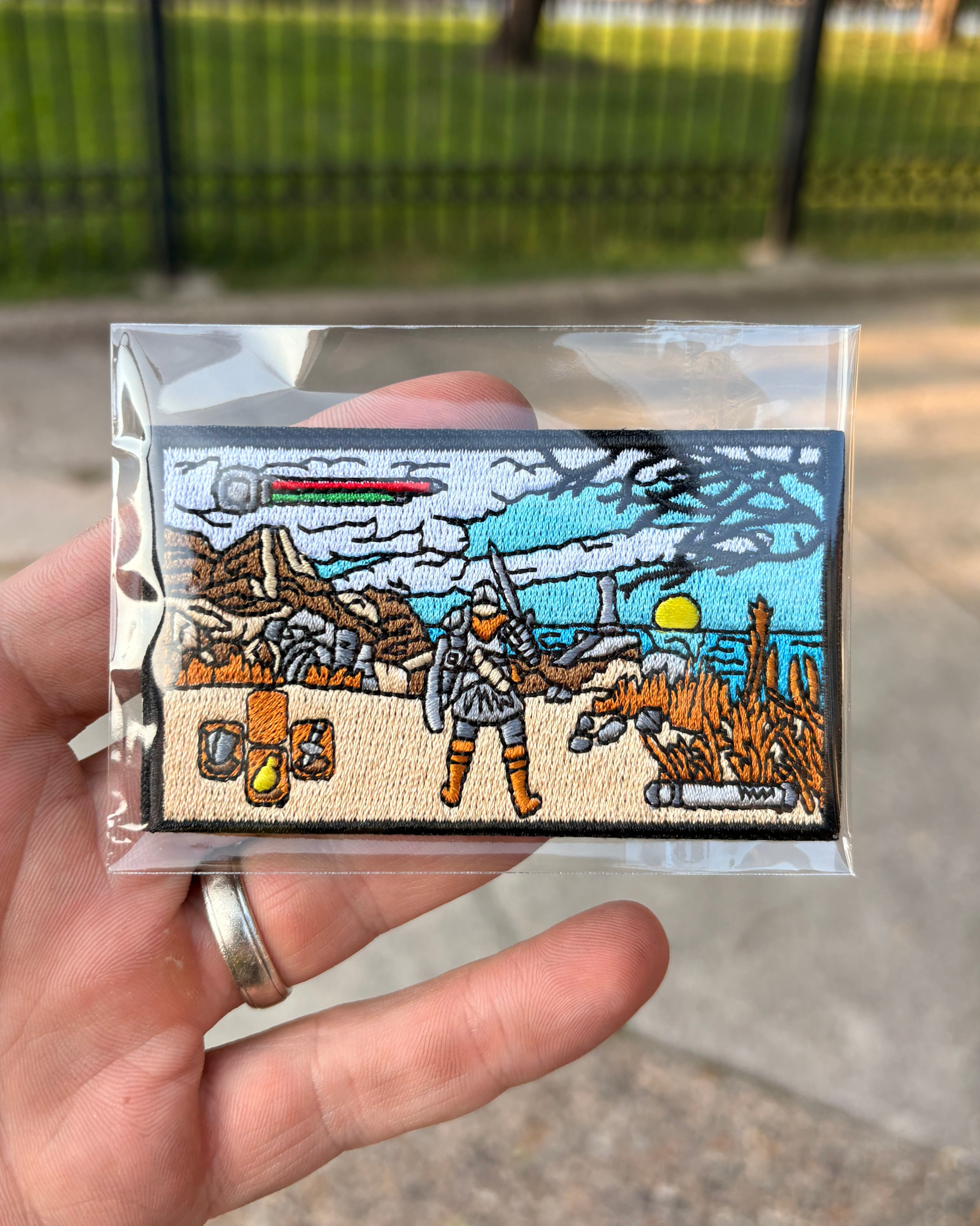 A hand holds a small embroidered patch inside a clear plastic sleeve, with a blurred outdoor background showing pavement and a fence. The patch depicts a sci-fi desert scene with a character in armor, rocks, plants, and a blue sky.