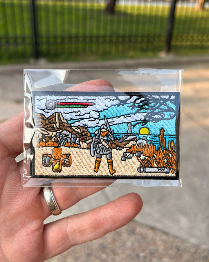 A hand holds a small embroidered patch inside a clear plastic sleeve, with a blurred outdoor background showing pavement and a fence. The patch depicts a sci-fi desert scene with a character in armor, rocks, plants, and a blue sky.