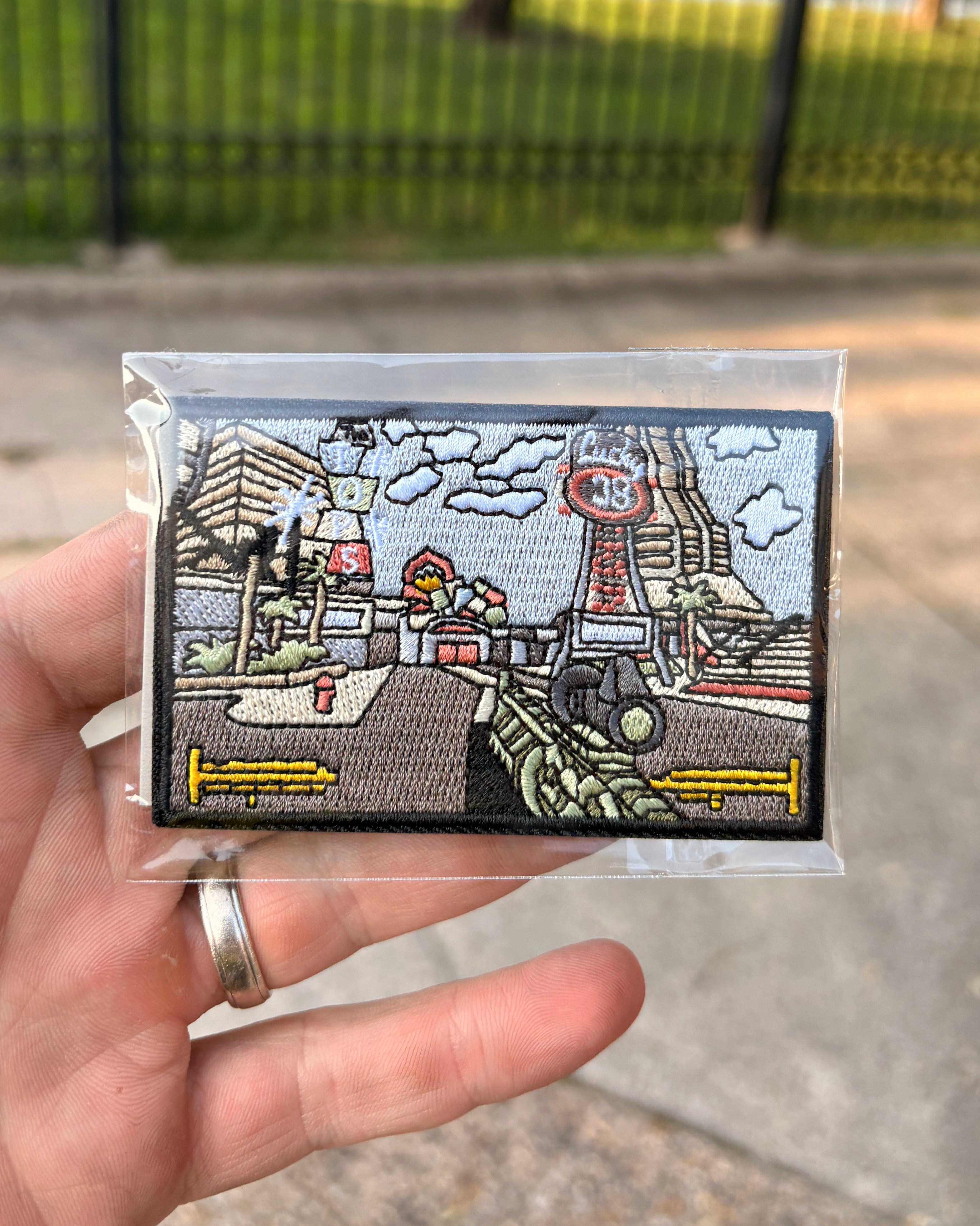 A hand holds a small rectangular embroidered patch inside a clear plastic sleeve, with an outdoor blurred background of pavement and a fence. The patch depicts a retro sci-fi city scene with a tower, clouds, and vehicles in gray, white, red, yellow, and black.