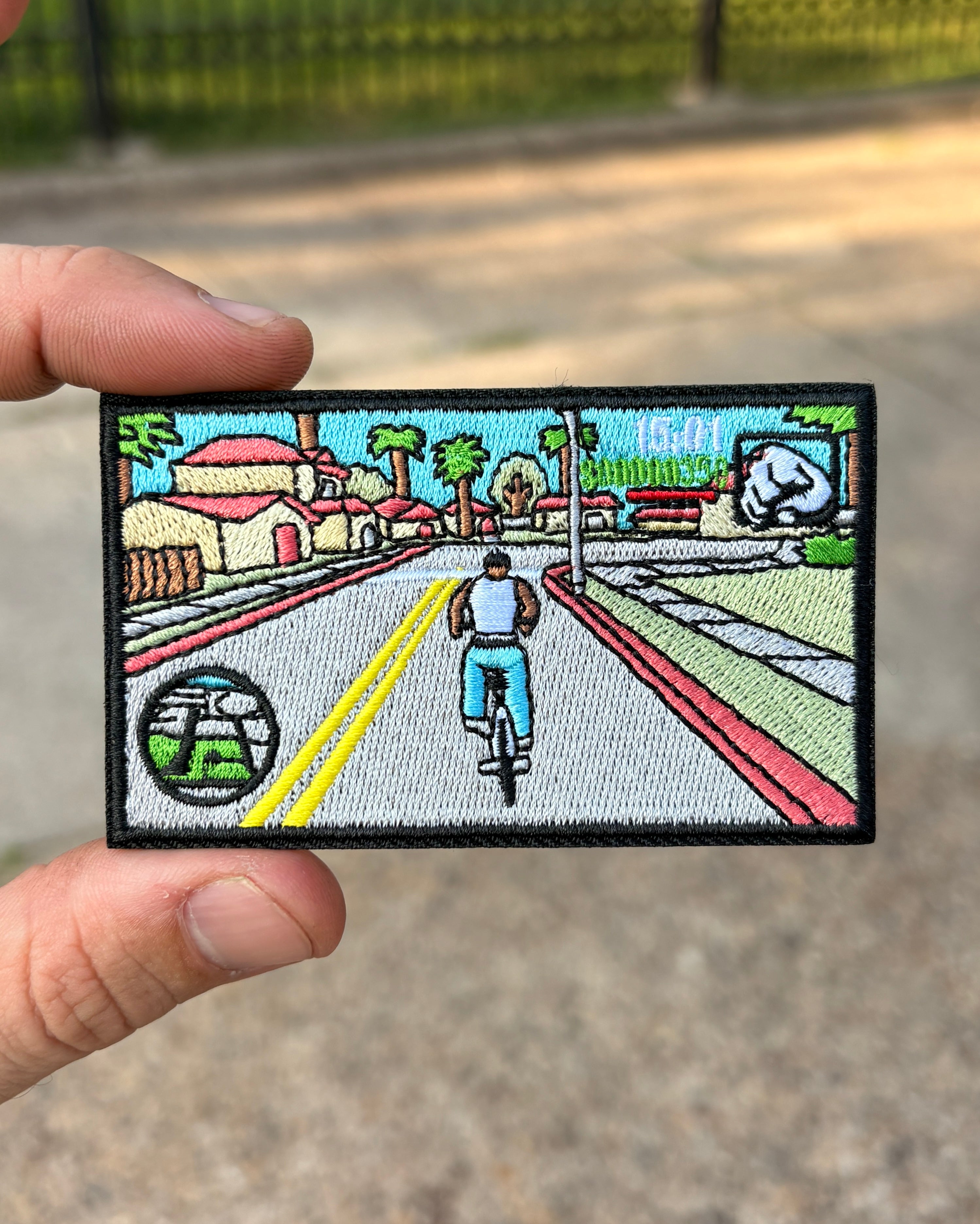 A rectangular embroidered patch held by a hand, featuring a retro-style illustration of a cyclist riding down a street with houses, trees, and a palm tree sign.