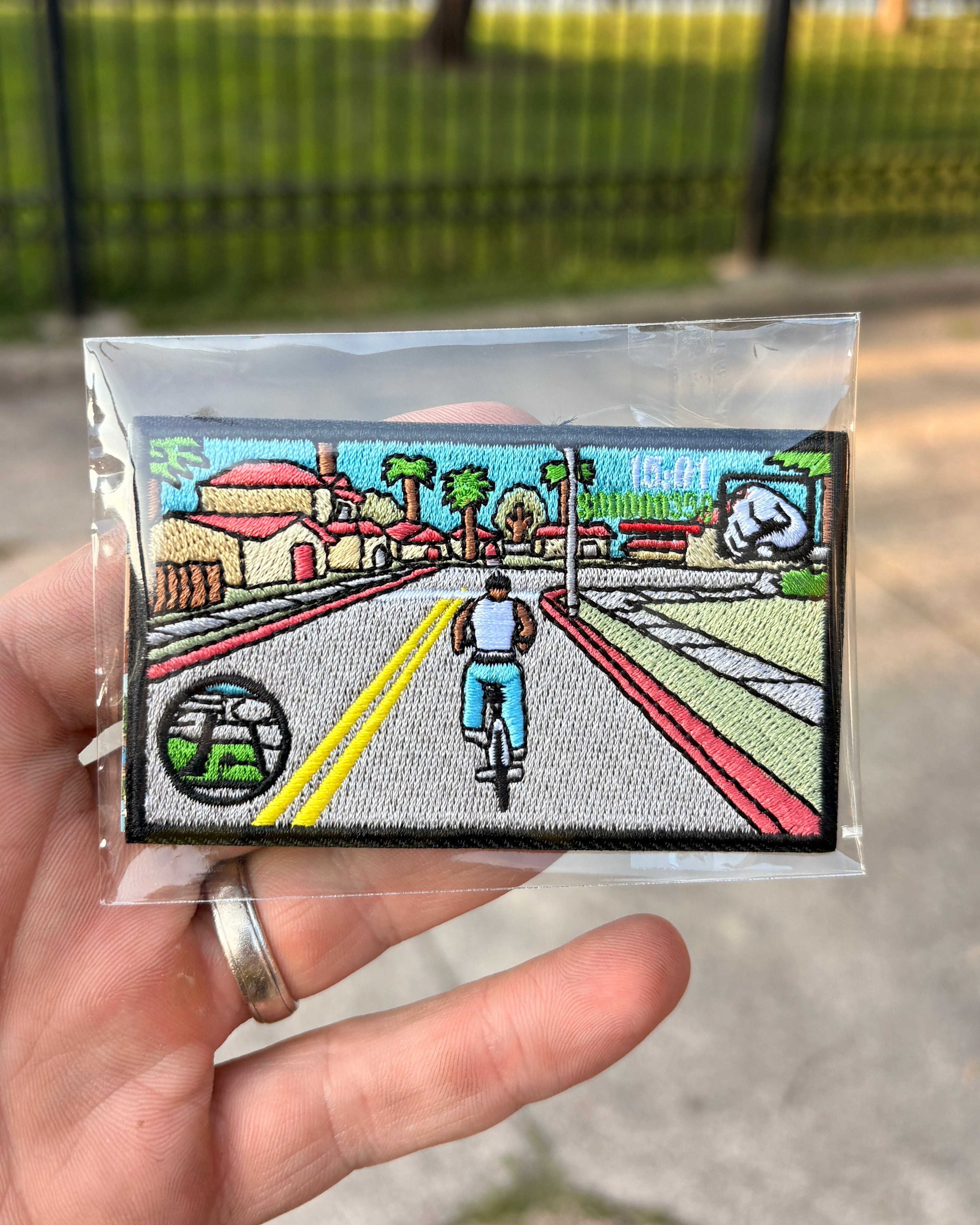 A hand holds a small embroidered patch inside a clear plastic sleeve, photographed outdoors with a blurred fence and pavement in the background. The patch depicts a cyclist riding down a street with houses, palm trees, a yellow double line, and a round emblem in the lower left.