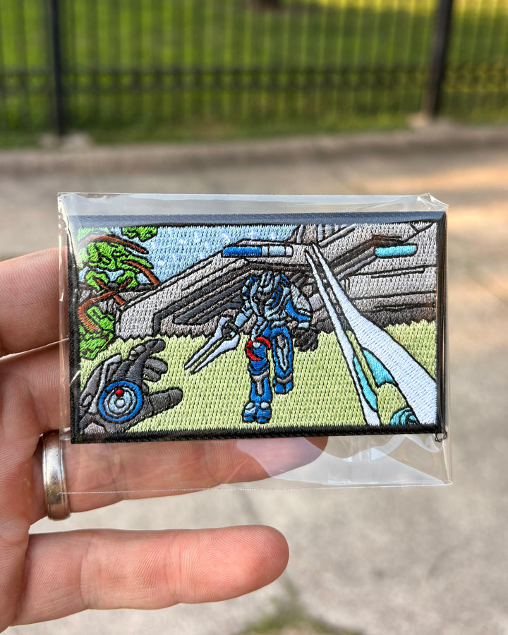 A hand holding a small embroidered patch inside a clear plastic sleeve, with an outdoor blurred background of pavement and a fence. The patch depicts a sci-fi scene with a blue robot and a spaceship.