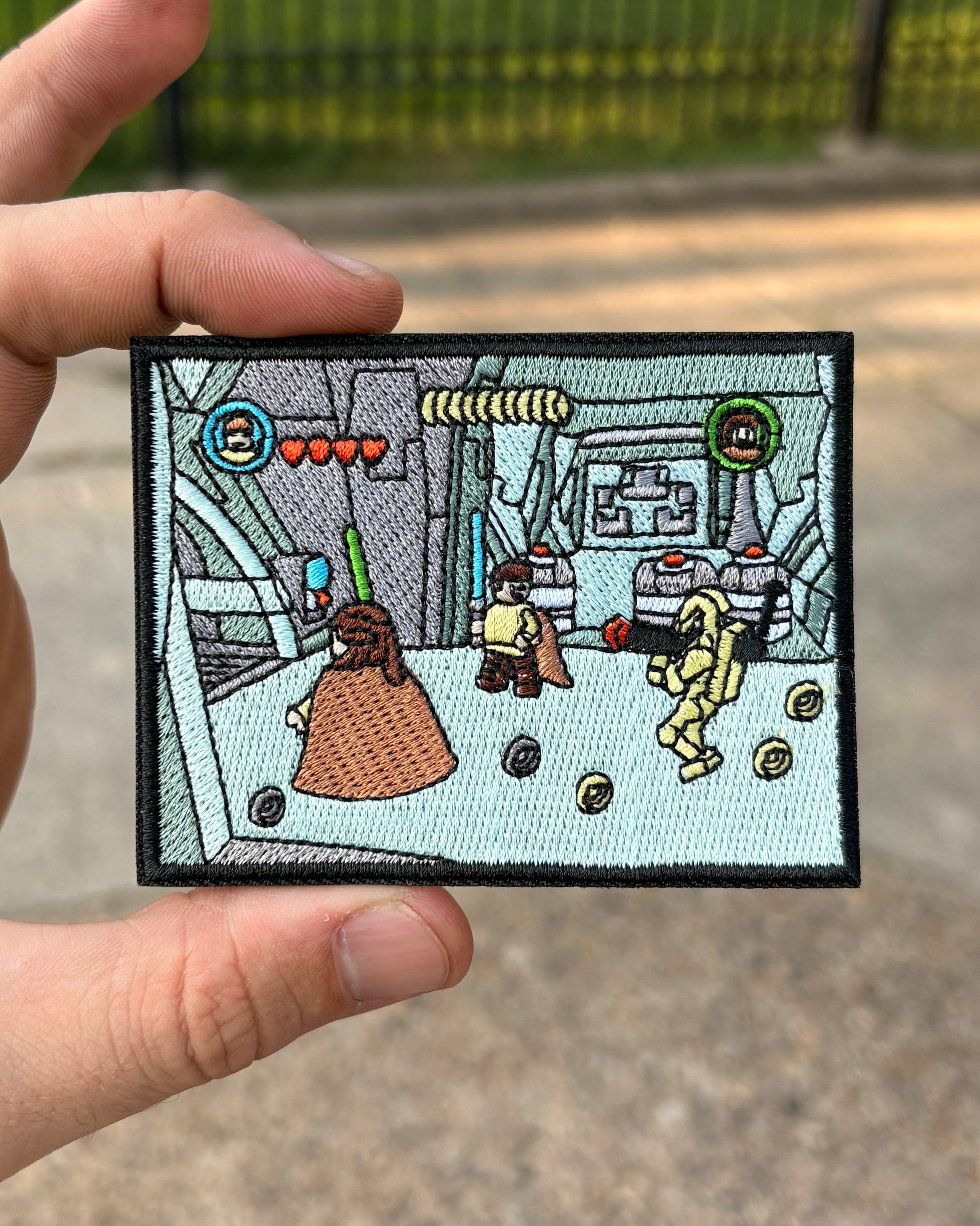 A hand holds a rectangular embroidered patch featuring a stylized sci-fi scene with small character figures, coins, and interface elements, against an outdoor blurred background.
