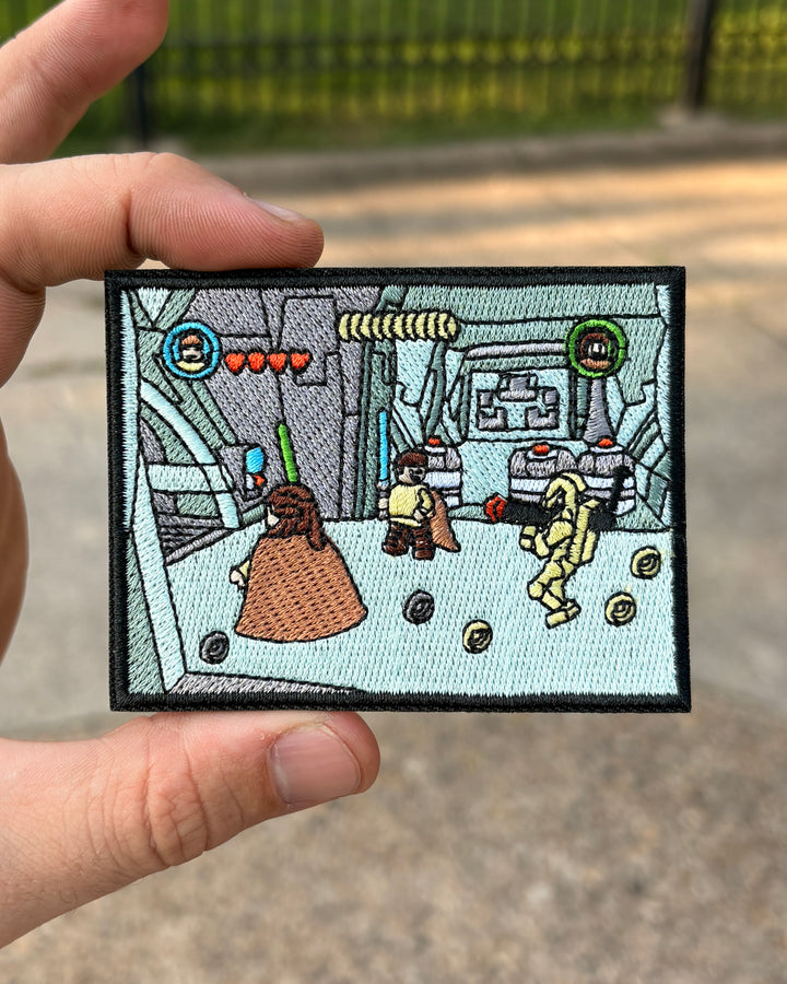 A hand holds a rectangular embroidered patch featuring a stylized sci-fi scene with small character figures, coins, and interface elements, against an outdoor blurred background.