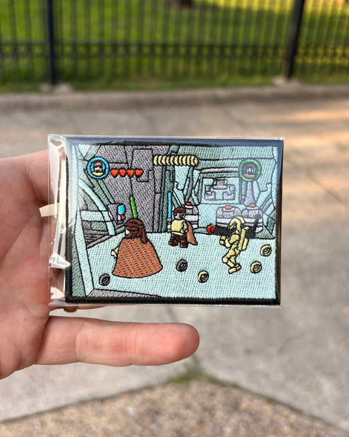 A hand holding a small rectangular embroidered patch featuring a pixel-art scene with characters and interface elements, photographed outdoors with a fence and pavement in the background.