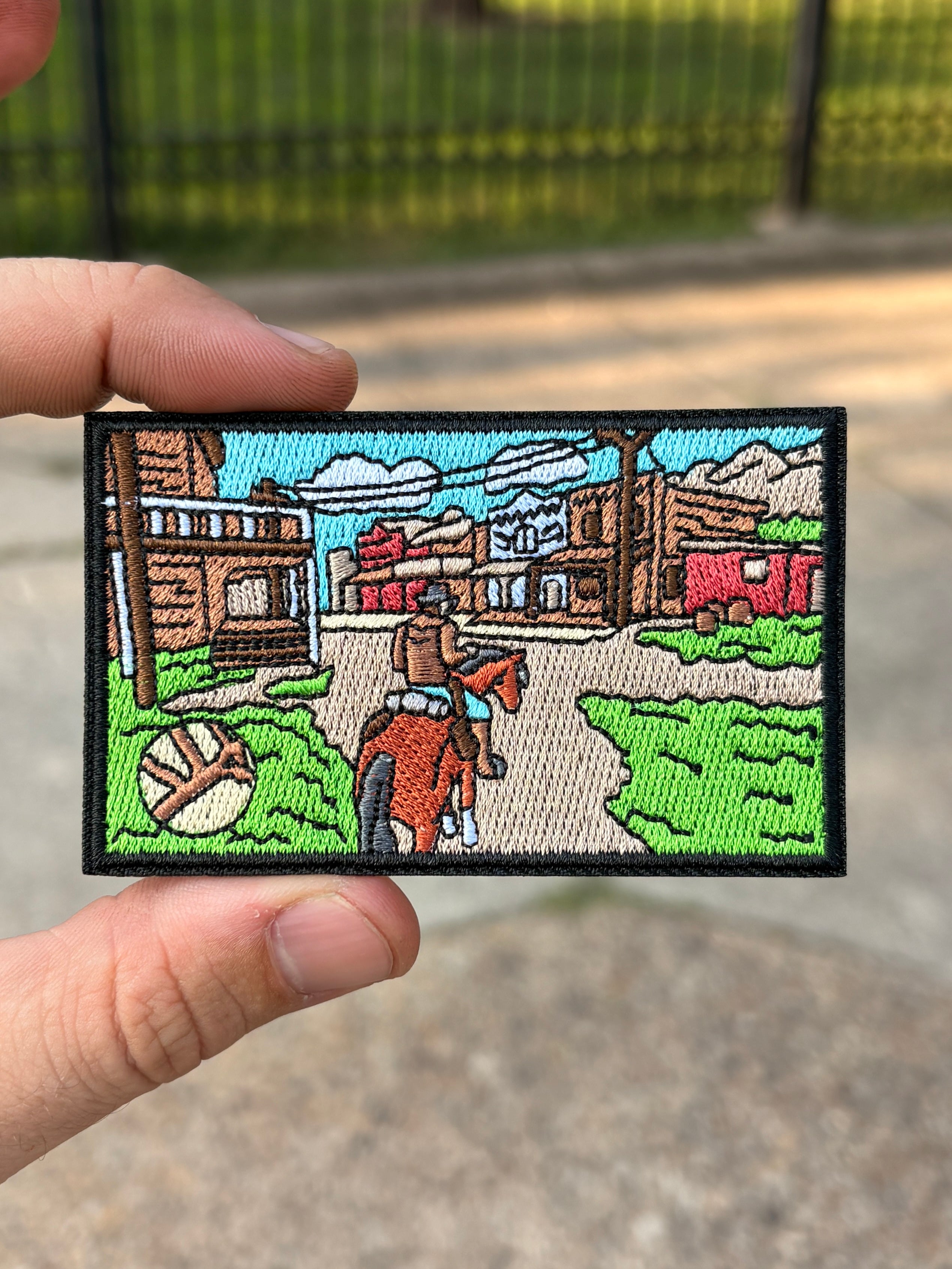 A rectangular embroidered patch held by two hands, featuring a western town scene with a cowboy on a horse, buildings, trees, and a blue sky.