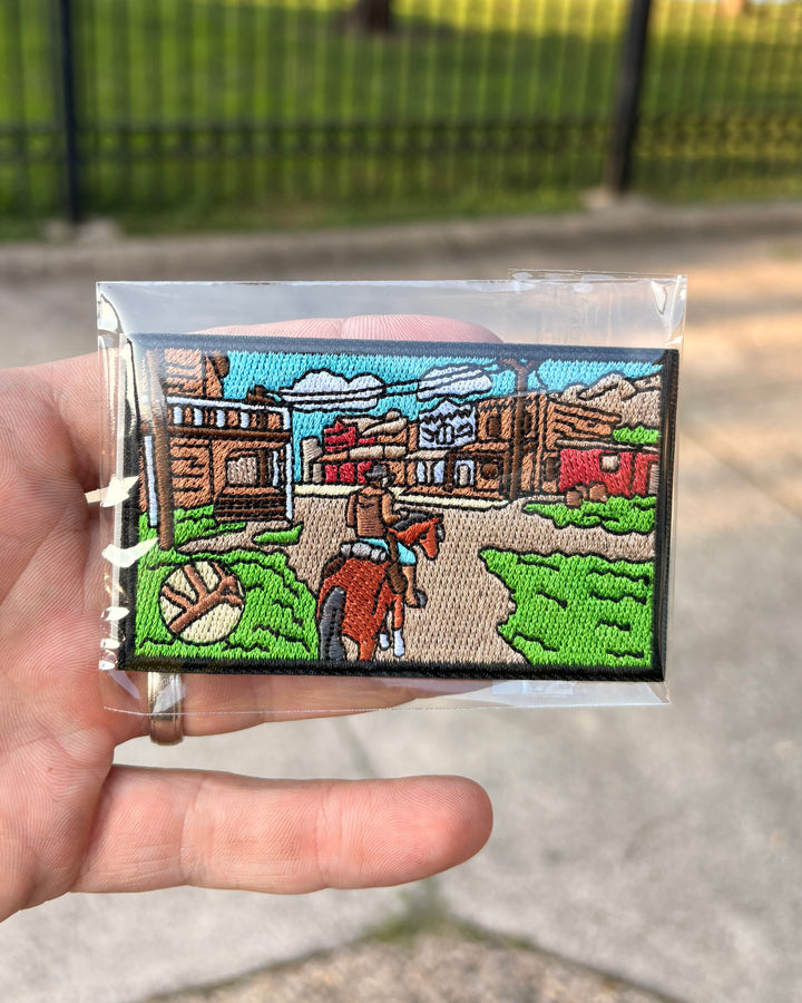 A hand holding a small embroidered patch in clear plastic packaging, with an outdoor blurred background of pavement and a fence. The patch depicts a western scene with a cowboy on a horse, buildings, trees, and a baseball.