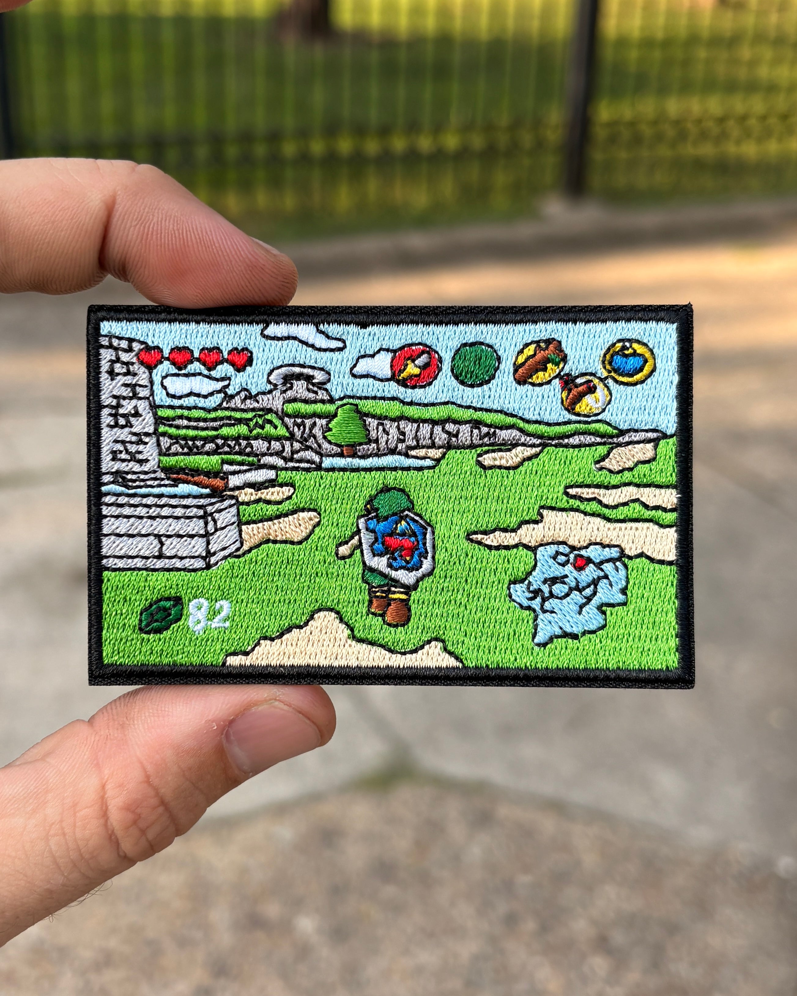A hand holds a rectangular embroidered patch featuring a colorful video game-style landscape with a character, clouds, and icons, against a blurred outdoor background.