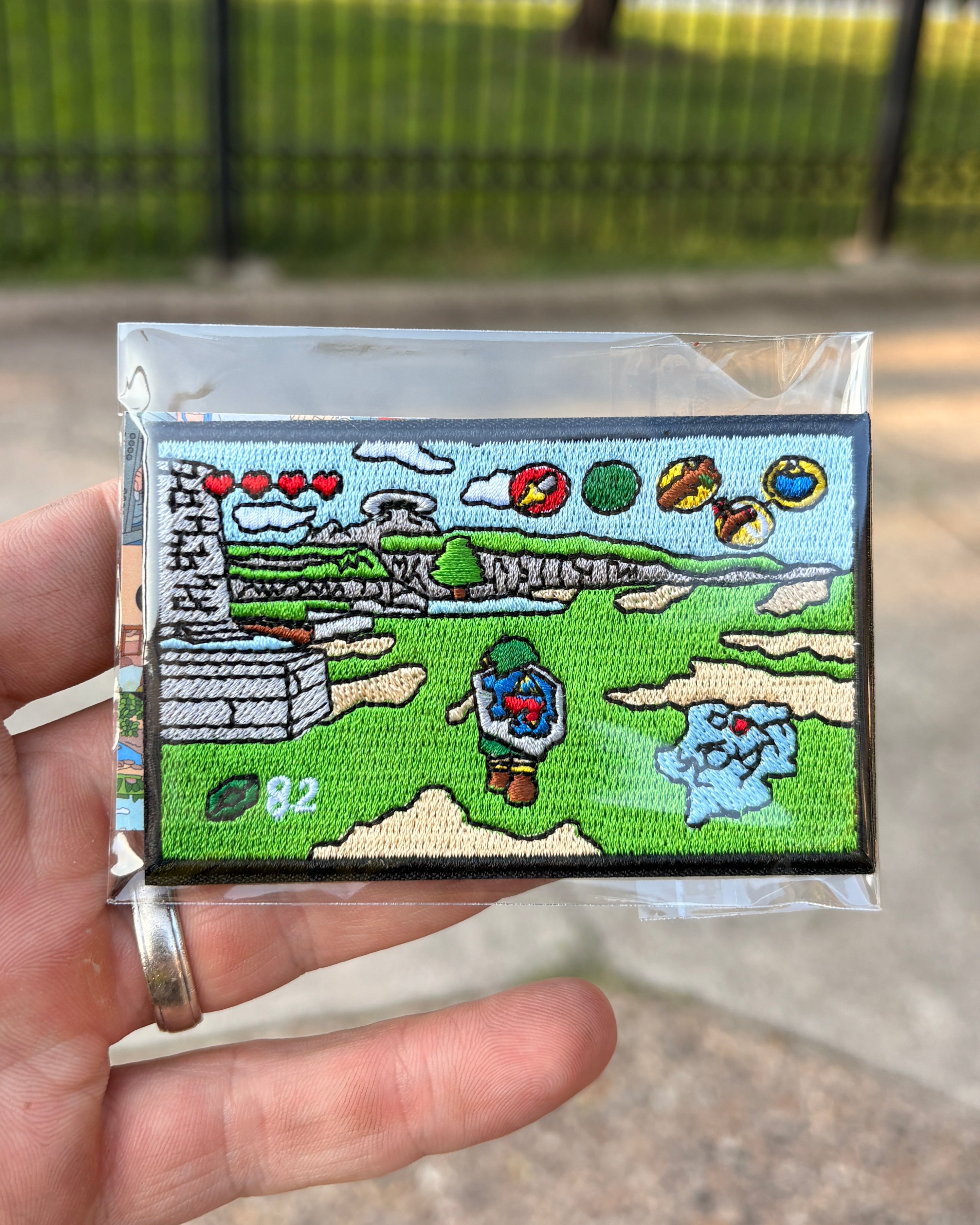 A hand holding a small embroidered patch inside a clear plastic sleeve, with a blurred outdoor background of grass and a fence.