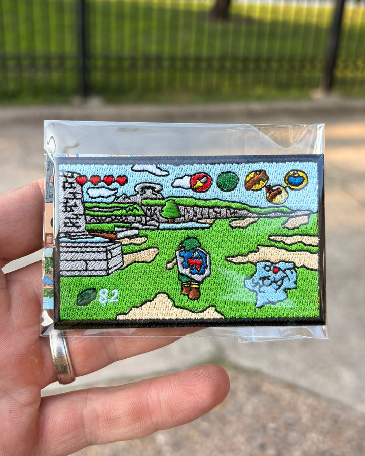 A hand holding a small embroidered patch inside a clear plastic sleeve, with a blurred outdoor background of grass and a fence.