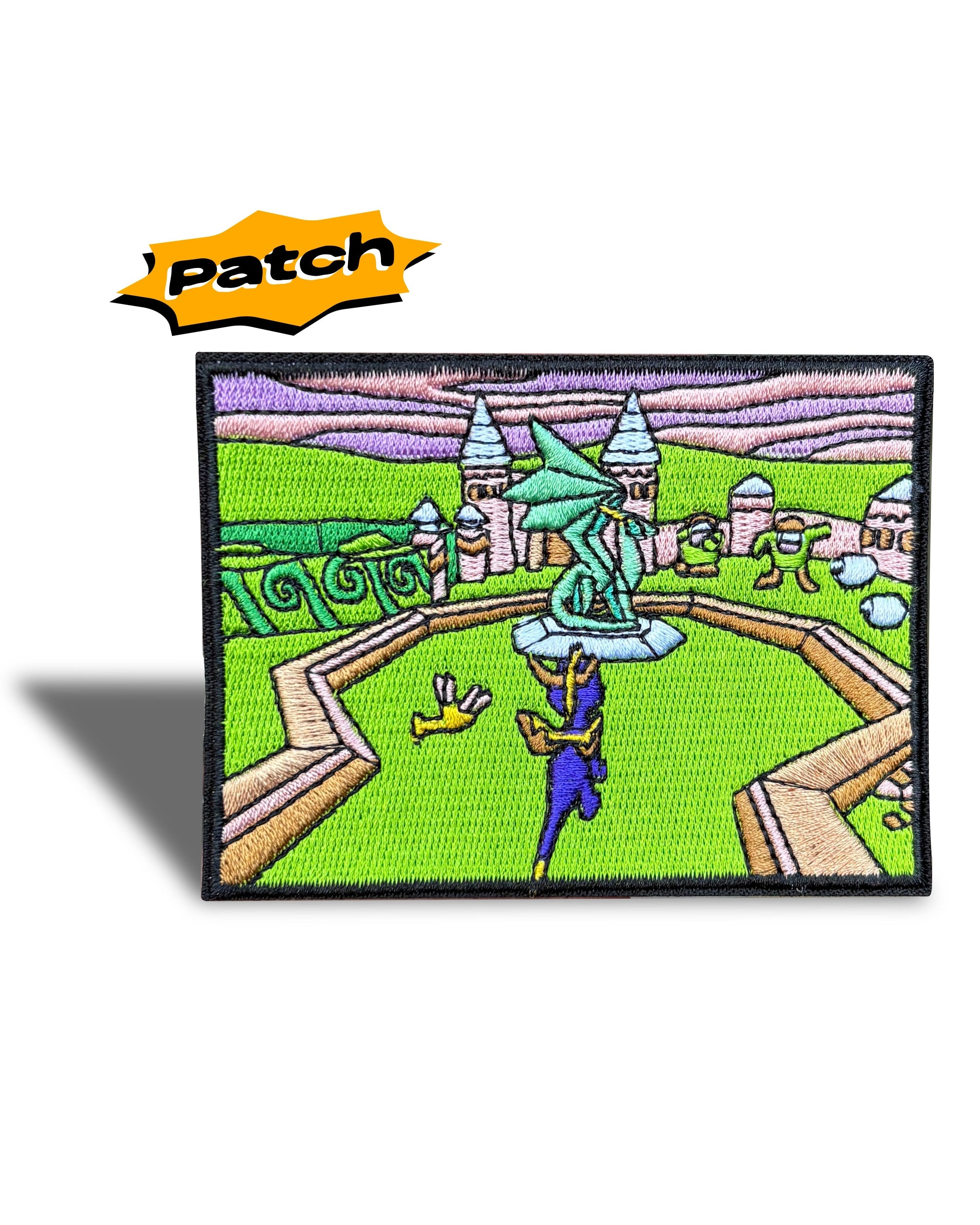 A rectangular embroidered patch featuring a colorful fantasy landscape with a green field, stone pathways, towers, and a blue-green statue, shown on a white background with a yellow 'patch' label above it.